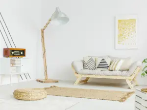 A modern living room with a light wood futon sofa, neutral patterned cushions, a tall floor lamp, woven rug, pouf, and wall art with yellow dots—perfect for enjoying stylish comfort in real estate properties Ulcinj Montenegro ekosphere.