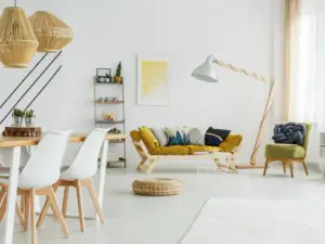 A modern living and dining area in Ulcinj, Montenegro, features white walls, wooden furniture, a yellow sofa with cushions, a green armchair, a floor lamp, decorative shelving, pendant lights, and a round woven rug—ideal for real estate properties seekers.