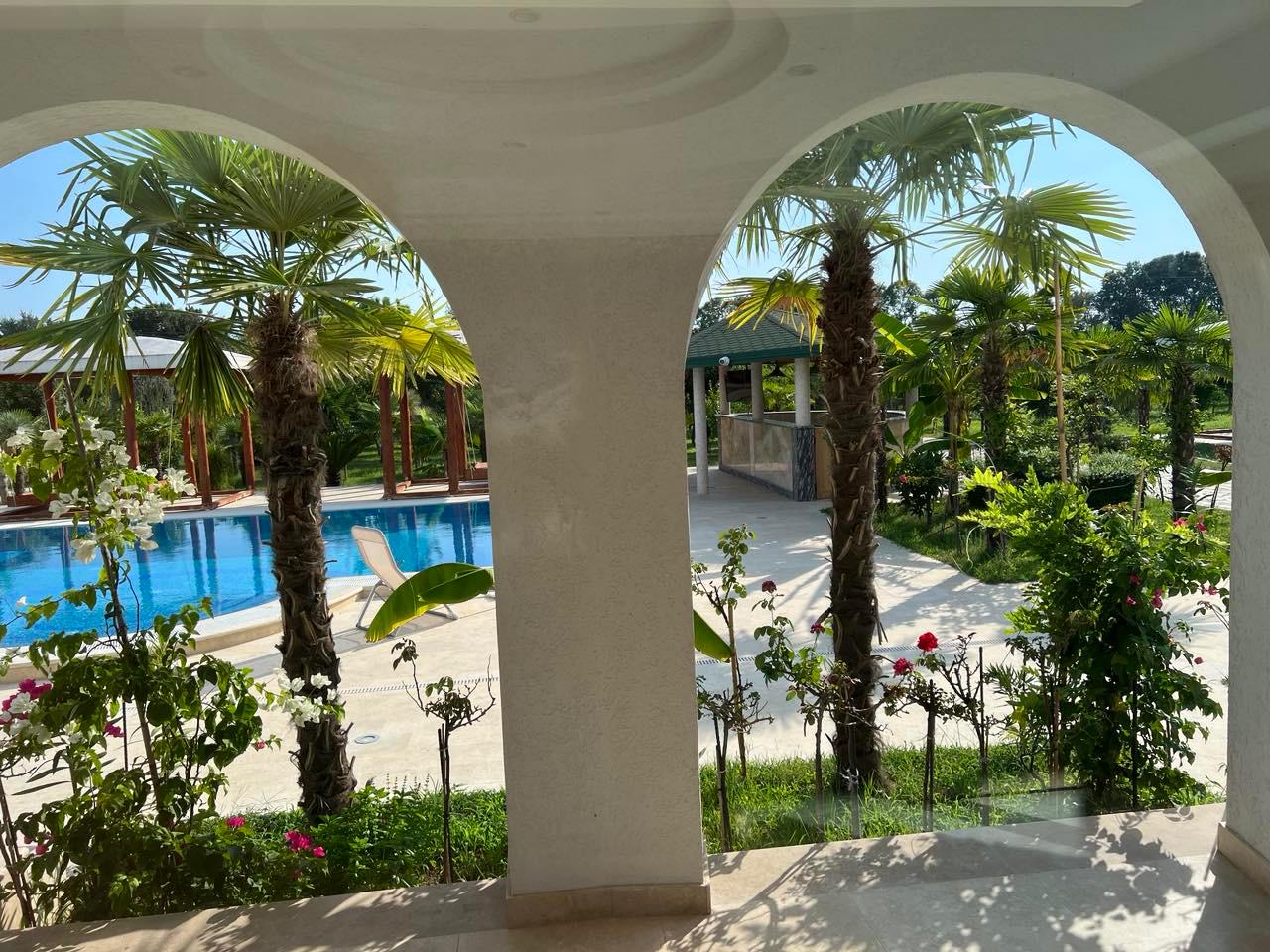 Stunning Ulcinj villa for sale: tropical garden, palm trees, pool view through white arches. Invest via montenegro-listings.me by Ekosphere Doo.