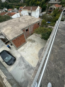 Balcony view of courtyard, black car, new brick construction & lush trees—prime Ulcinj investment. Listed on montenegro-listings.me by ekosphere doo.