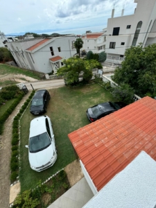 Aerial view of Ulcinj real estate: red-roofed home, grassy lot with two cars, white houses nearby—your investment at montenegro-listings.me by ekosphere doo.