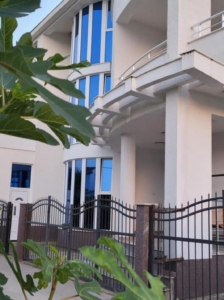 Modern multi-story white building with blue-tinted windows, lush greenery, and secure fencing—prime Ulcinj property on montenegro-listings.me by ekosphere doo.