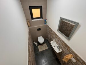 Modern small bathroom in Ulcinj property, with toilet, wall sink, mirror, and window. Invest via montenegro-listings.me by ekosphere doo.