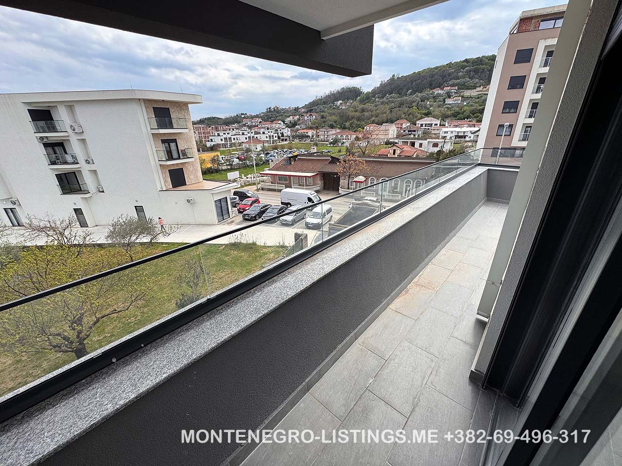 Stunning balcony view with glass railing over prime Ulcinj real estate&mdash;buildings, cars, lush hills. montenegro-listings.me by Ekosphere doo.
