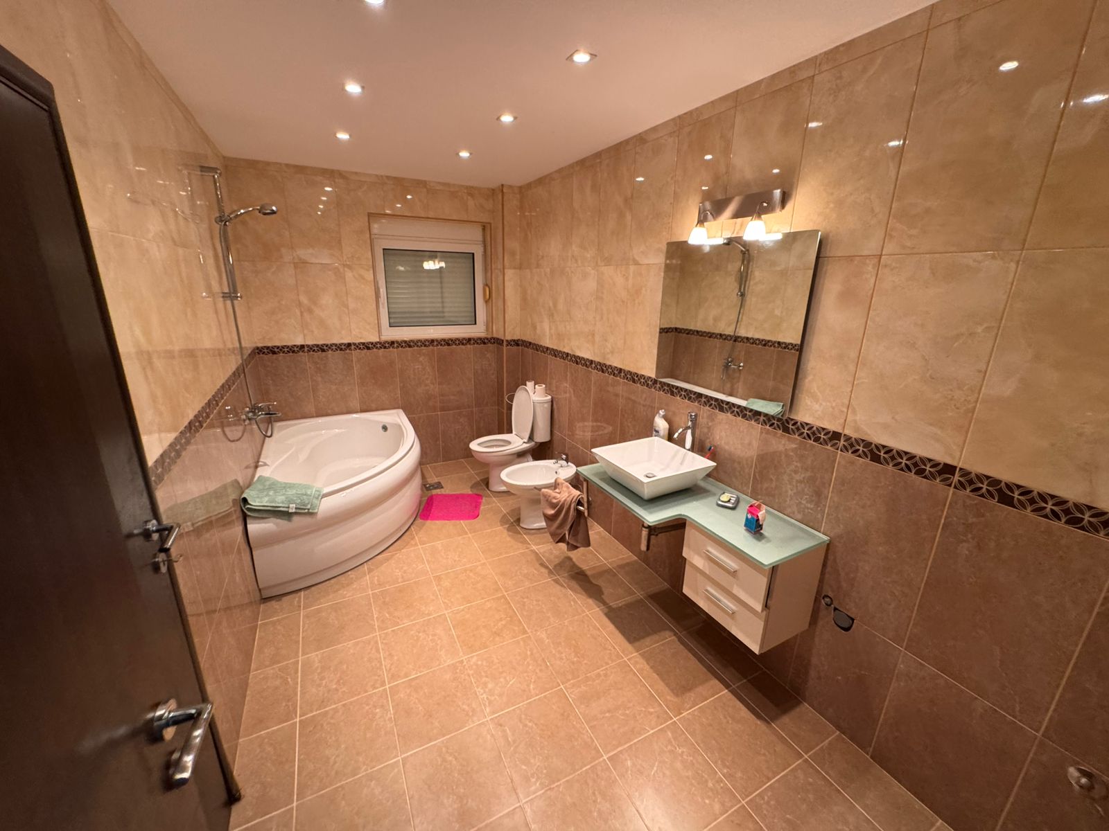 Modern bathroom in Ulcinj with beige tiles, corner tub, window, and quality fixtures—exclusive property at montenegro-listings.me by ekosphere doo.