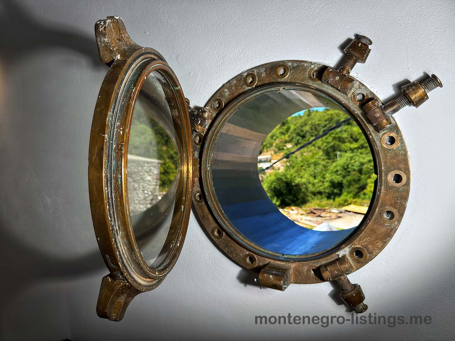Open brass porthole on white wall reveals lush Ulcinj views&mdash;explore top Montenegro real estate at montenegro-listings.me by ekosphere doo.