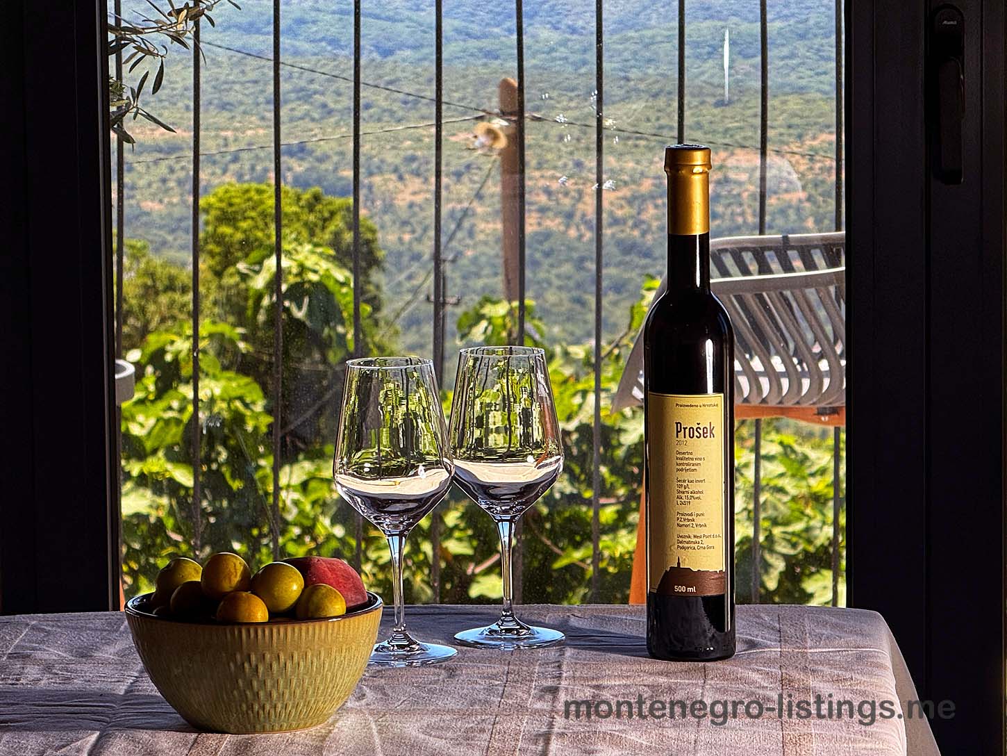 Pro&scaron;ek wine, glasses, and fresh fruit set by a window with green hill views&mdash;experience Ulcinj real estate luxury with montenegro-listings.me.