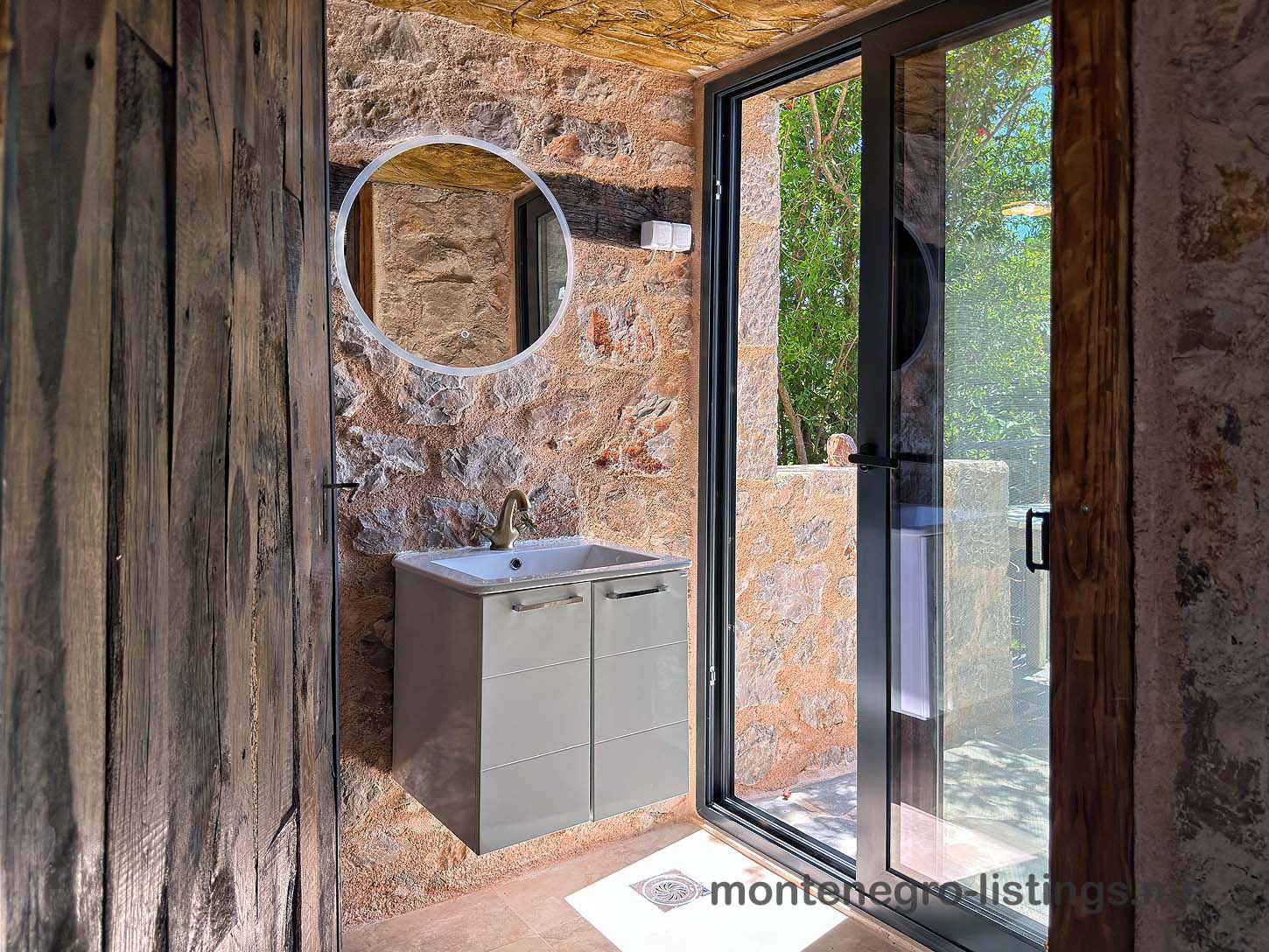 Luxury modern bathroom in Ulcinj, Montenegro: elegant gray vanity, stone walls, garden view. Invest with montenegro-listings.me by Ekosphere.