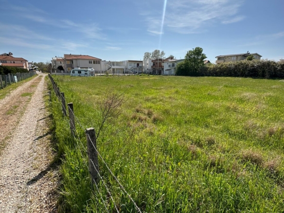 Prime Ulcinj investment: Spacious grassy field with wire fence and gravel access, near sunny homes. Listed by ekosphere doo.