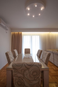 Elegant Ulcinj dining area: glass table, designer chairs, sunlit windows, chic lighting, hardwood floors. Invest via montenegro-listings.me by ekosphere doo.