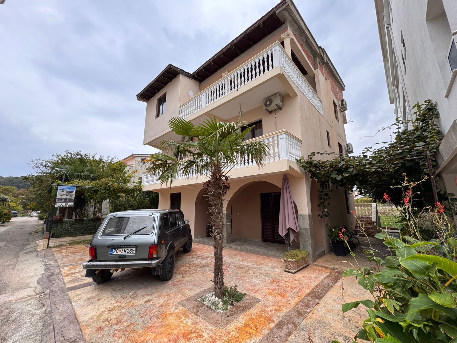Elegant three-story beige villa with balconies, lush greenery, palm tree, and private driveway in Ulcinj. Listed on montenegro-listings.me by ekosphere doo—prime real estate investment opportunity!.