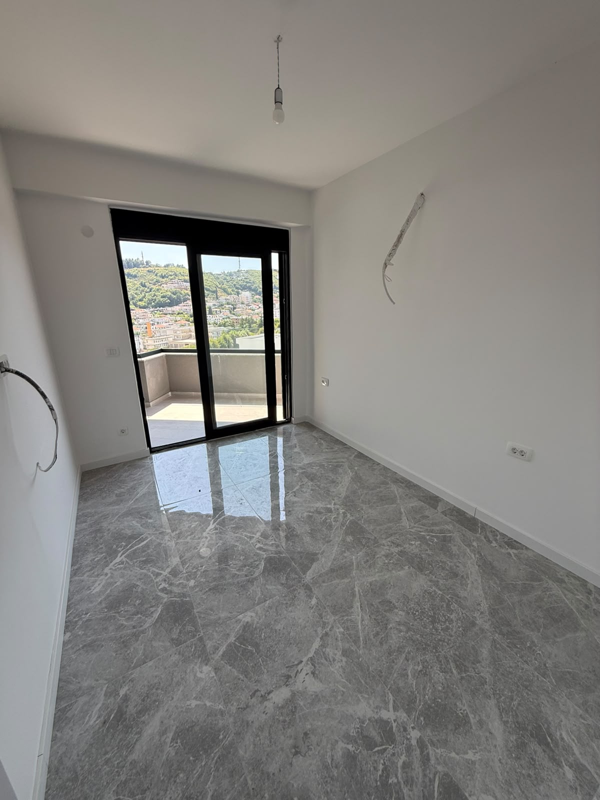 Spacious, sunlit room with premium gray marble flooring and crisp white walls—an ideal canvas for your dream home or investment property in Ulcinj, Montenegro. Expansive glass doors welcome natural light and provide seamless access to a private balcony with panoramic city views and lush greenery, perfect for relaxing or entertaining guests. This property presents a unique opportunity for those seeking to invest in the thriving Montenegro real estate market through montenegro-listings.me, a trusted project of ekosphere doo in Ulcinj. Modern touches await your finishing details, making this listing an exceptional choice for buyers and investors alike.