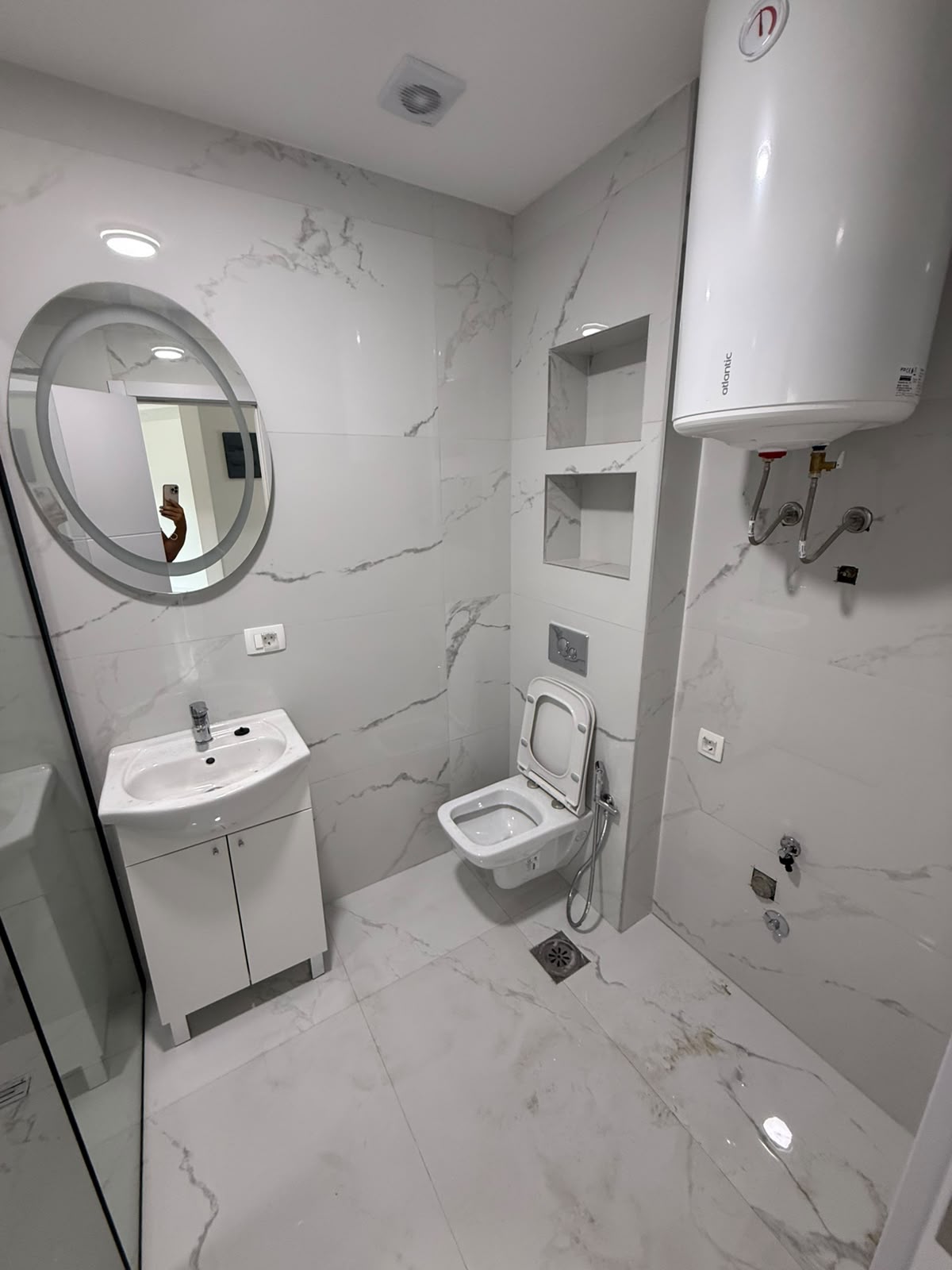 Discover a stylish, modern bathroom featuring elegant white marble tiles in this prime Ulcinj real estate opportunity. Enjoy sophisticated amenities such as a sleek round mirror above the contemporary sink, a wall-mounted toilet for optimal space and comfort, state-of-the-art electric water heating, and thoughtfully designed built-in shelving for ultimate functionality. This clean and minimally furnished space is perfect for investors seeking high-value property in Montenegro’s fast-growing market. Presented by montenegro-listings.me—a project of ekosphere doo in Ulcinj—this property combines luxury finishes with profitable investment potential on the Adriatic coast.