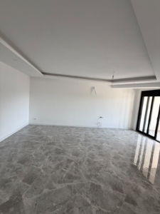 Bright and spacious empty room in Ulcinj, Montenegro, featuring pristine white walls, elegant gray marble flooring, and stylish black-framed sliding glass doors that invite abundant natural light. The modern white ceiling with recessed lighting adds to the airy ambiance, while conveniently placed electrical outlets and cabling offer practical flexibility. This well-lit property offers exceptional potential for investors or homebuyers seeking premium real estate in Ulcinj. Explore more high-value opportunities on montenegro-listings.me, a trusted project by ekosphere doo in Ulcinj.