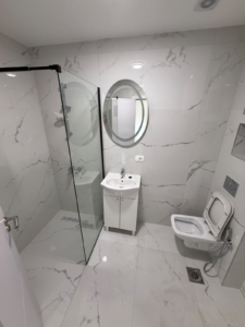 Experience luxury living in this contemporary bathroom, featuring elegant white marble walls and flooring that exude sophistication. The space is enhanced by a sleek glass shower enclosure, a stylish oval mirror above a modern sink with built-in cabinet storage, and a space-saving wall-mounted toilet. Perfectly suited for discerning buyers seeking prime real estate investment opportunities in Ulcinj, Montenegro, this exceptional property is listed exclusively on montenegro-listings.me—a trusted project by ekosphere doo in Ulcinj. Discover the perfect blend of comfort and value for your next investment in the thriving Montenegro property market.