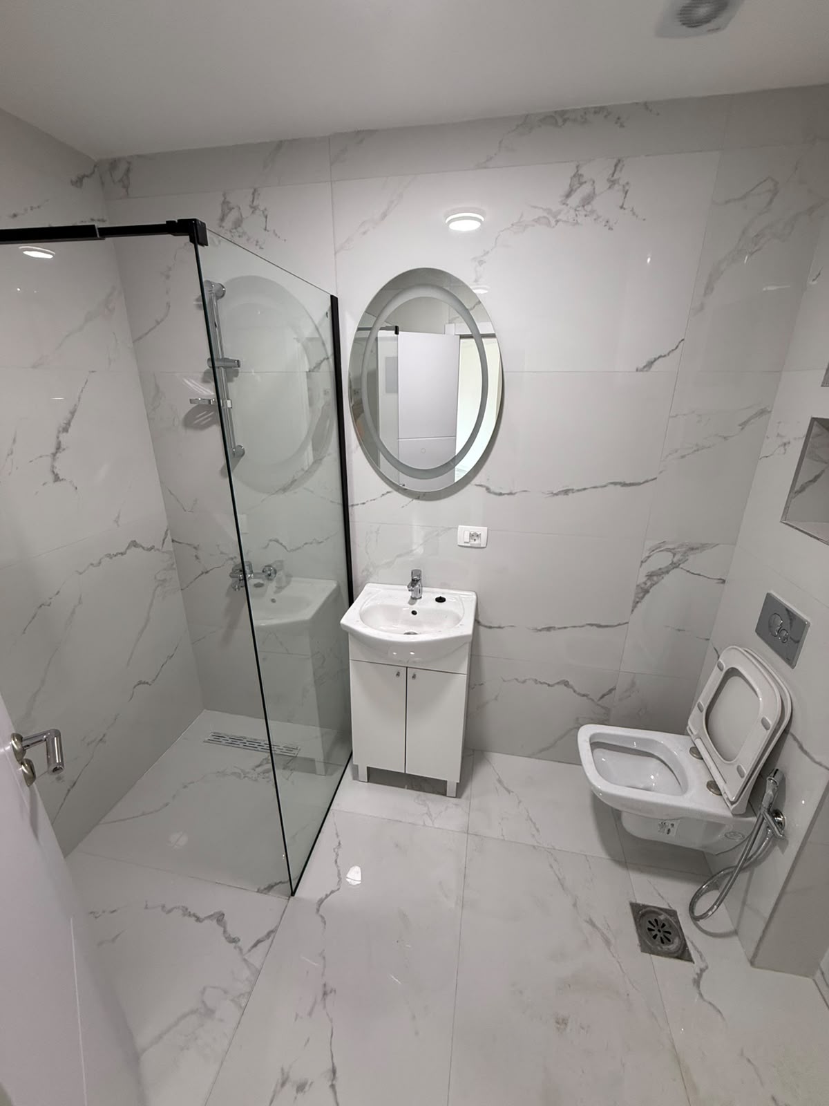 Experience luxury living in this contemporary bathroom, featuring elegant white marble walls and flooring that exude sophistication. The space is enhanced by a sleek glass shower enclosure, a stylish oval mirror above a modern sink with built-in cabinet storage, and a space-saving wall-mounted toilet. Perfectly suited for discerning buyers seeking prime real estate investment opportunities in Ulcinj, Montenegro, this exceptional property is listed exclusively on montenegro-listings.me—a trusted project by ekosphere doo in Ulcinj. Discover the perfect blend of comfort and value for your next investment in the thriving Montenegro property market.