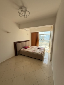 Bright and inviting bedroom in Ulcinj, Montenegro, perfect for property investment or summer living. This well-lit room features a comfortable double bed positioned against the left wall, accented with plush pillows and blanket for immediate coziness. A large window with an elegant orange curtain fills the space with natural sunlight, highlighting the pristine white walls and stylish tiled floors—offering a fresh, modern feel. Ideal for buyers seeking high-potential Montenegro real estate or Ulcinj holiday homes. Available exclusively through montenegro-listings.me—a trusted project of ekosphere doo in Ulcinj—this property merges comfort with exceptional investment opportunity.