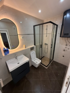 Experience contemporary living in this stylish Ulcinj, Montenegro bathroom, featuring a sleek lit oval mirror above a modern sink, pristine white toilet, elegant black-framed glass shower enclosure, and energy-efficient water heater. Perfect for discerning property buyers and investors seeking top-quality real estate in Montenegro. Discover exceptional investment opportunities with montenegro-listings.me—a trusted project of ekosphere doo in Ulcinj—offering sophisticated finishes and premium amenities tailored to the Montenegro property market.