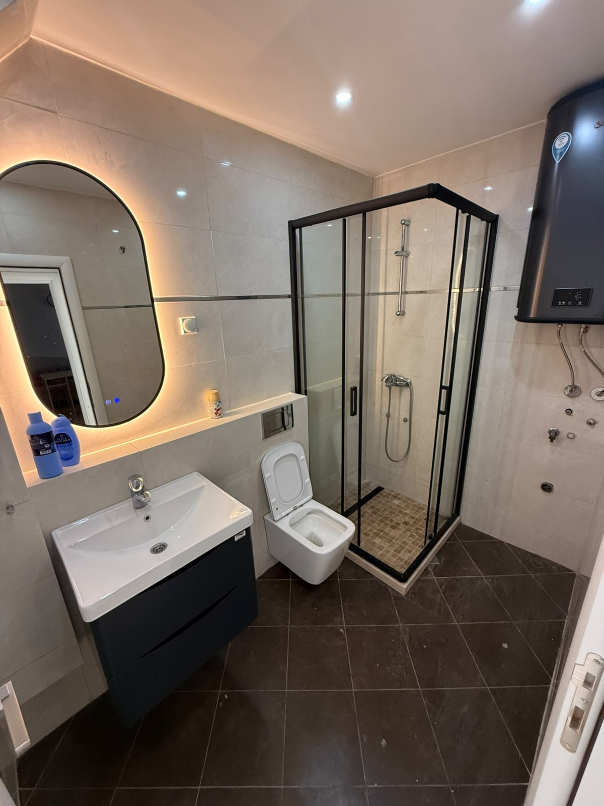 Experience contemporary living in this stylish Ulcinj, Montenegro bathroom, featuring a sleek lit oval mirror above a modern sink, pristine white toilet, elegant black-framed glass shower enclosure, and energy-efficient water heater. Perfect for discerning property buyers and investors seeking top-quality real estate in Montenegro. Discover exceptional investment opportunities with montenegro-listings.me—a trusted project of ekosphere doo in Ulcinj—offering sophisticated finishes and premium amenities tailored to the Montenegro property market.