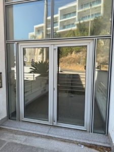 Modern glass door with a sleek metal frame, reflecting an inviting outdoor space featuring stone steps, elegant railing, lush greenery, and a neighboring building—perfect for those seeking premium real estate in Ulcinj, Montenegro. This property showcases quality craftsmanship and versatile indoor-outdoor living potential, ideal for investment or your new Mediterranean home. Explore exclusive Ulcinj listings on montenegro-listings.me—a trusted project by ekosphere doo—to secure your place in Montenegro’s thriving property market.