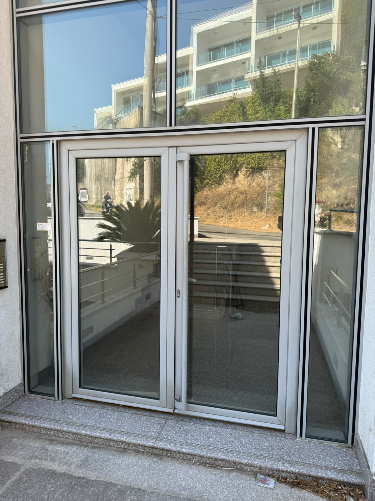 Modern glass door with a sleek metal frame, reflecting an inviting outdoor space featuring stone steps, elegant railing, lush greenery, and a neighboring building—perfect for those seeking premium real estate in Ulcinj, Montenegro. This property showcases quality craftsmanship and versatile indoor-outdoor living potential, ideal for investment or your new Mediterranean home. Explore exclusive Ulcinj listings on montenegro-listings.me—a trusted project by ekosphere doo—to secure your place in Montenegro’s thriving property market.