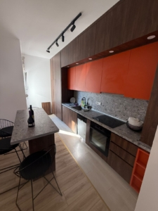 Experience contemporary living in this exquisite Ulcinj, Montenegro kitchen, featuring sleek brown and orange cabinetry, a stylish gray stone countertop, and premium built-in appliances—perfect for investment or family living. The spacious island with elegant black wire chairs creates an inviting atmosphere for dining or entertaining, complemented by modern track lighting overhead. This turnkey property opportunity is showcased on montenegro-listings.me, a project of ekosphere doo in Ulcinj—a trusted name in Montenegrin real estate.