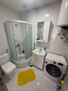 Modern, fully-equipped small bathroom featuring pristine white tiles, a sleek glass shower enclosure, contemporary toilet, and a stylish sink with integrated storage cabinet. Includes additional wall cabinet for optimal organization and an in-unit washing machine topped with fresh folded towels for everyday convenience. Thoughtfully accented by a cheerful yellow rug and neatly arranged toiletries. This inviting space enhances property value—an ideal investment opportunity in Ulcinj, Montenegro’s booming real estate market. For exclusive listings by ekosphere doo in Ulcinj, visit montenegro-listings.me.