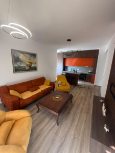 Experience contemporary coastal living in this stylish Ulcinj property, exclusively listed on montenegro-listings.me—a project of ekosphere doo. The spacious open-plan living room features vibrant orange and yellow seating, a sleek wooden coffee table, and a wall-mounted TV—perfect for relaxing or entertaining guests. The modern kitchen boasts striking red cabinets, ideal for culinary enthusiasts. Nautical-inspired artwork and elegant decorative lighting add sophisticated touches to this inviting space. Invest in premium Montenegro real estate with excellent rental potential right at the heart of Ulcinj’s thriving property market.