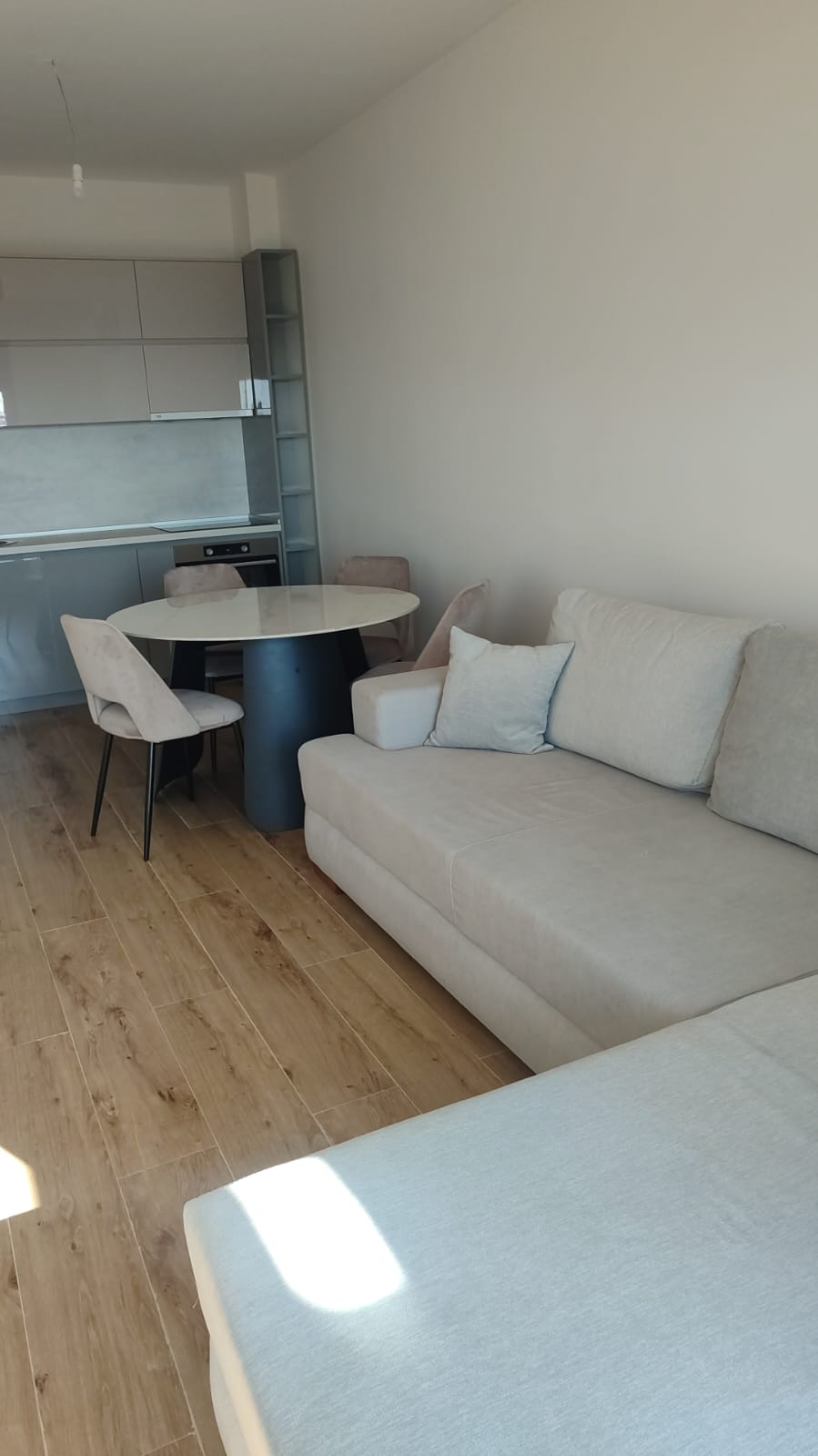 Discover a stylish modern living space in Ulcinj, Montenegro—an exceptional real estate investment opportunity featured on montenegro-listings.me, a project by ekosphere doo. This impeccably designed apartment showcases elegant light wood floors, a spacious beige sectional sofa perfect for relaxing or entertaining guests, and a contemporary round dining table with seating for four. The sleek compact kitchen is fitted with premium gray cabinets and complements the open-plan layout. Abundant natural light flows in from the left side, creating an inviting and airy atmosphere ideal for year-round living or holiday rentals in Montenegro’s bustling property market.