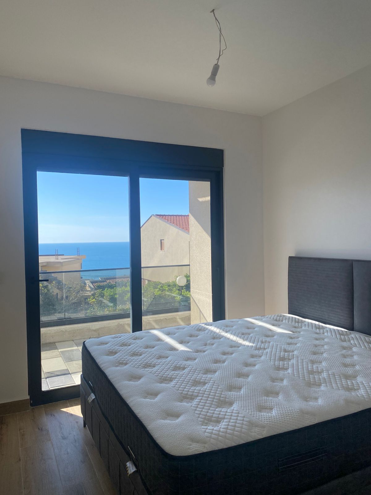 Spacious Ulcinj bedroom featuring a modern gray headboard, quality hardwood flooring, and expansive glass doors that open to a private balcony with breathtaking views of the Adriatic Sea, picturesque rooftops, and clear Montenegrin skies. Natural light floods the room, creating an inviting investment opportunity in the heart of Montenegro’s coastal property market. Discover premium real estate through montenegro-listings.me—a trusted project by ekosphere doo in Ulcinj—perfect for buyers seeking ocean-view homes or vacation rentals.