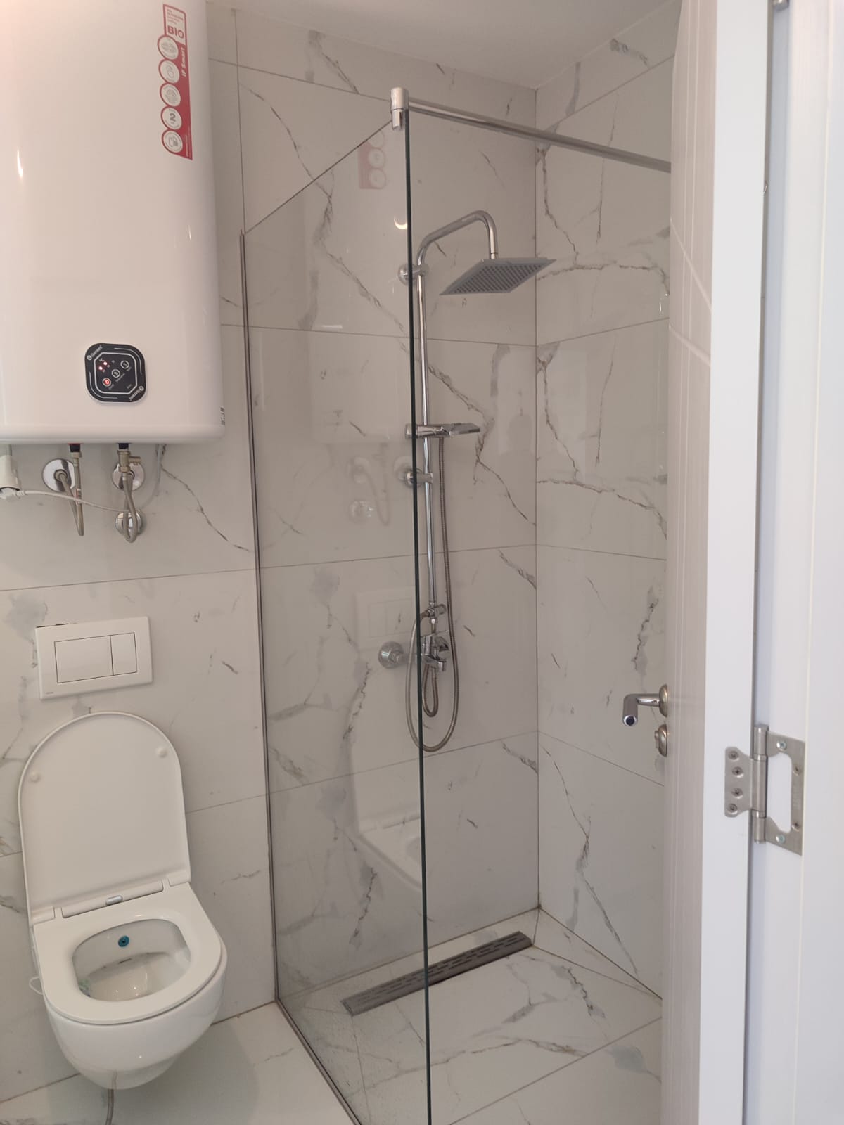 Elegant bathroom in Ulcinj, Montenegro, featuring a modern wall-mounted toilet, luxurious glass-enclosed shower with a rainfall showerhead, and stylish marble-patterned tiles, complemented by an efficient white water heater above the toilet. This high-quality finish offers both comfort and functionality—ideal for real estate investors seeking premium property opportunities in the sought-after Ulcinj coastal market. Discover exceptional investments at montenegro-listings.me, a project proudly presented by ekosphere doo in Ulcinj.