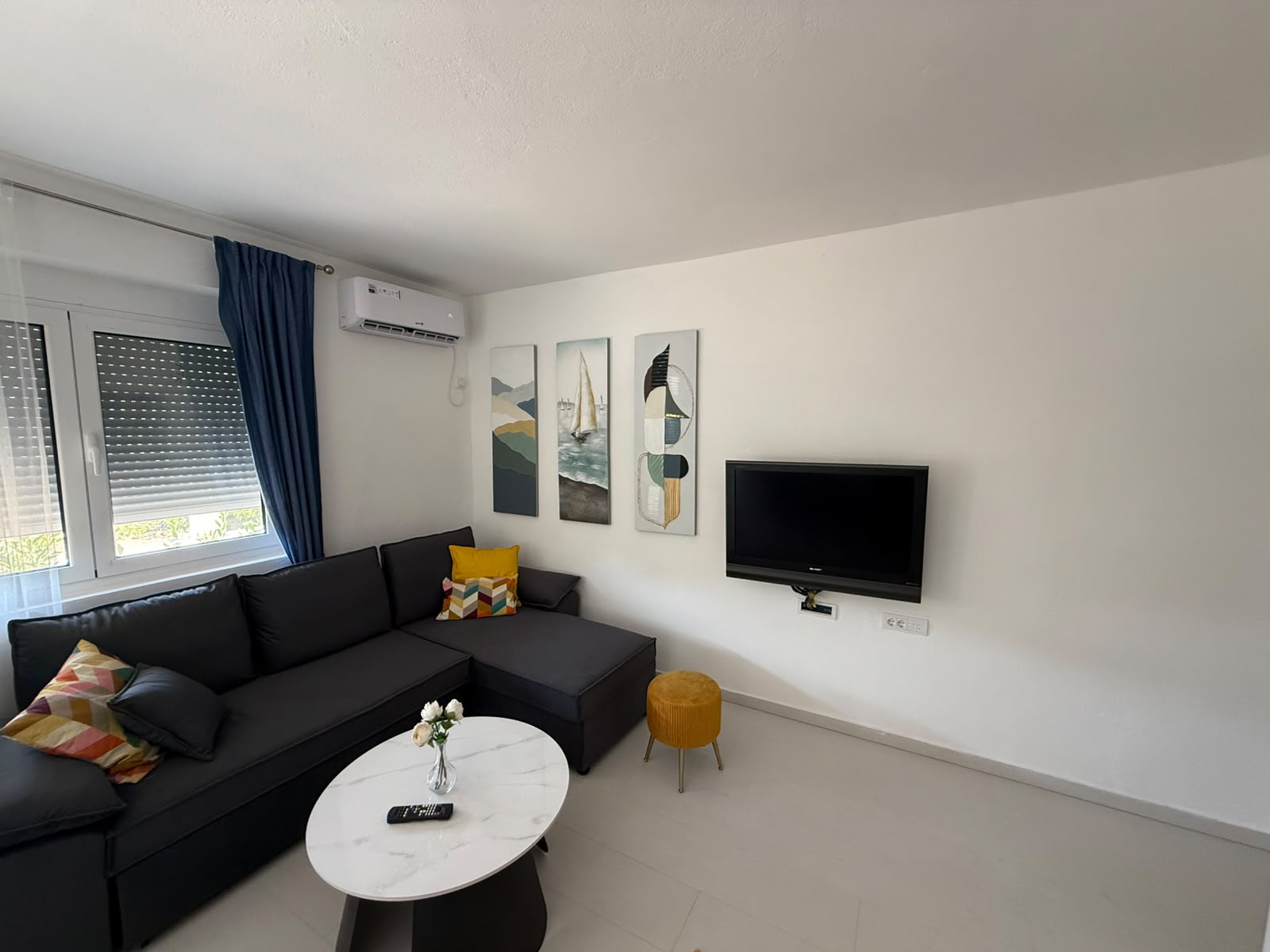 Discover this stylish, contemporary living room in Ulcinj, Montenegro—an ideal real estate investment showcased exclusively on montenegro-listings.me, a project of ekosphere doo. The spacious interior features a sophisticated dark L-shaped sofa with vibrant accent pillows, complemented by a sleek white oval coffee table and a chic yellow ottoman. Nautical-themed wall art adds coastal charm, while the wall-mounted TV and large window with elegant blue curtains and a modern roller blind elevate comfort and relaxation. Perfectly suited for property buyers seeking premium homes or investment opportunities in the booming Montenegro property market.