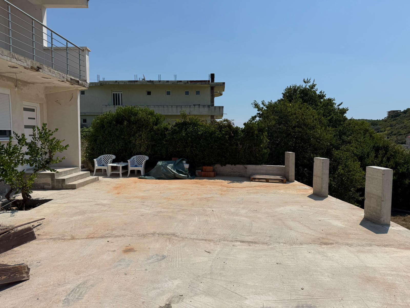 Spacious concrete patio ideal for outdoor entertaining, featuring four white plastic chairs and ample space for gatherings. This inviting terrace is adjacent to a well-maintained white building and offers serene views of lush greenery and distant architecture under a bright, clear blue sky. Perfectly situated in Ulcinj, Montenegro, this property presents excellent investment potential in the thriving Montenegrin real estate market. Discover more exclusive opportunities at montenegro-listings.me, a project proudly developed by ekosphere doo in Ulcinj.