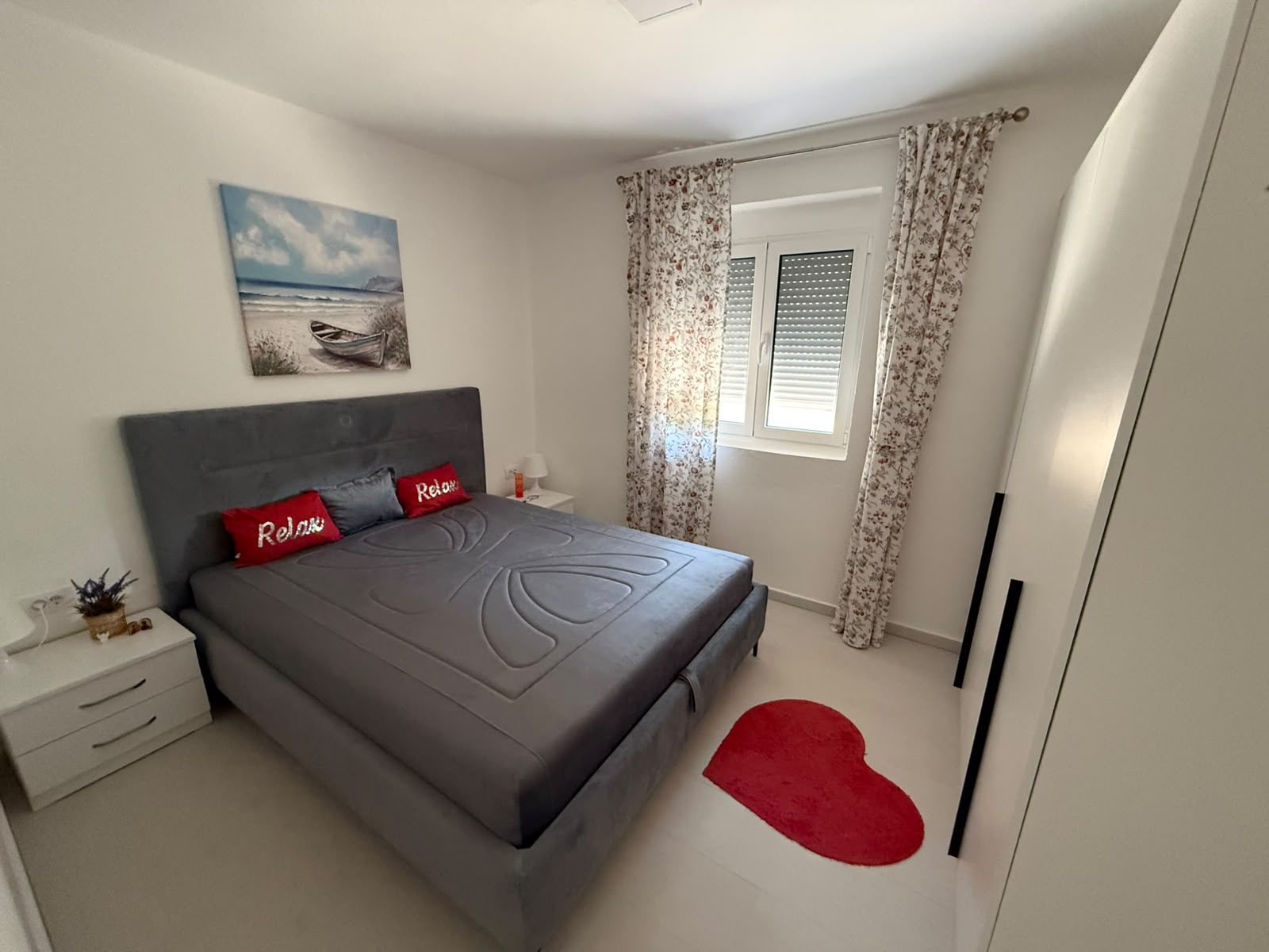Experience comfortable coastal living in this beautifully appointed bedroom in Ulcinj, Montenegro. The space features a stylish gray bed accented by two red “Relax” pillows, complemented by modern white nightstands and elegant floral curtains. Enjoy natural light from the window with closed blinds, while the unique red heart-shaped rug adds a touch of charm. Above the bed, a captivating seascape painting enhances the tranquil ambiance. This inviting property is an excellent investment opportunity in the Montenegro real estate market—discover more exclusive listings at montenegro-listings.me, a project of ekosphere doo based in Ulcinj.