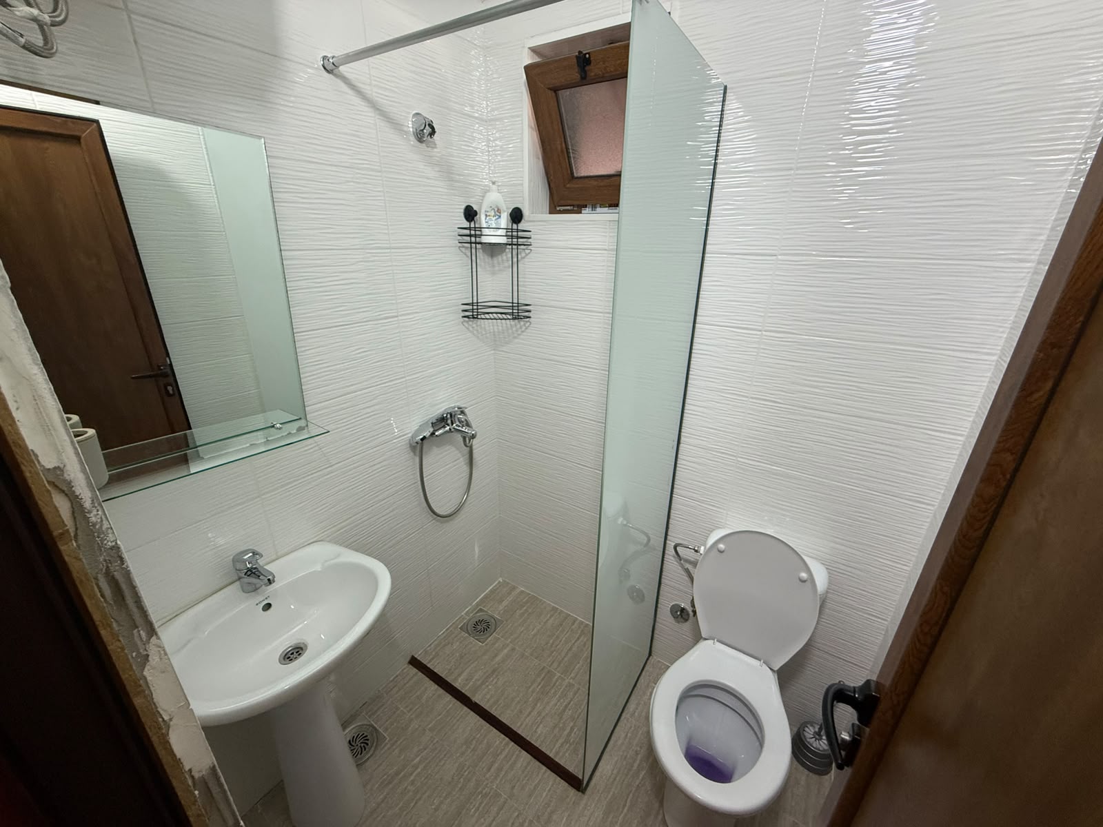 Bright, modern white bathroom featuring premium fixtures including a sleek pedestal sink, wall-mounted mirror, and contemporary toilet—perfect for sophisticated living in Ulcinj, Montenegro. The spacious corner glass shower is complemented by a convenient metal rack for toiletries and natural light from the window above. This thoughtfully designed space combines style and functionality, making it an ideal investment opportunity listed exclusively on montenegro-listings.me—a premier project by ekosphere doo in Ulcinj—highlighting the finest real estate options in the Montenegro property market.