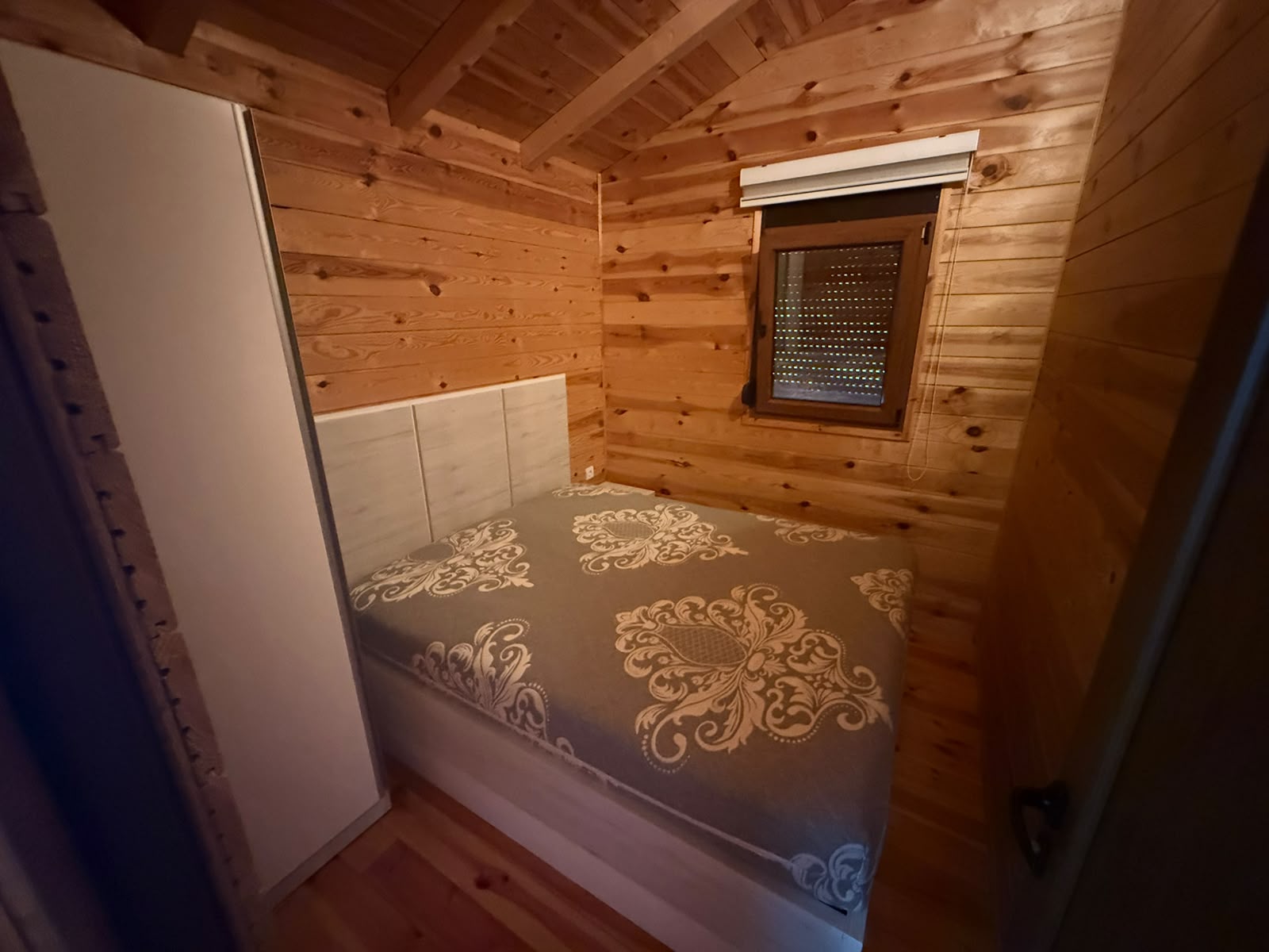 Experience cozy living in this charming wooden bedroom, featuring a comfortable double bed with an elegant gray patterned bedspread, complemented by a sleek white wardrobe and a bright window with privacy blind. The walls and ceiling are crafted from premium light-colored wood, creating a warm and inviting atmosphere that is perfect for year-round comfort. This meticulously maintained space is an ideal investment opportunity in the heart of Ulcinj, Montenegro—whether for personal use or as a lucrative rental property. Discover value and quality with properties like this on montenegro-listings.me, brought to you by ekosphere doo Ulcinj—your trusted local real estate partner.