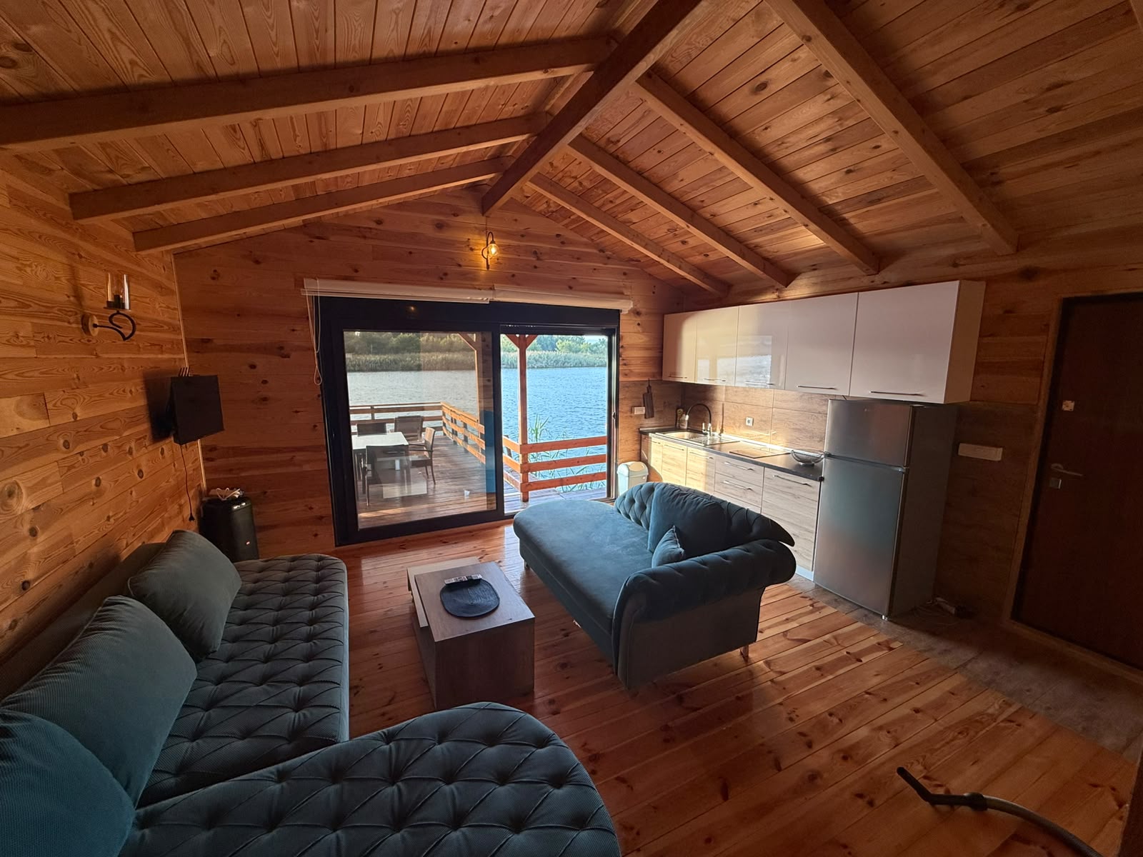 Experience authentic lakefront living in Ulcinj, Montenegro with this stunning wooden cabin retreat, offered exclusively through montenegro-listings.me—a project of ekosphere doo. The inviting interior showcases two stylish blue sofas, a modern kitchenette with refrigerator, and a cozy dining area—perfect for relaxing or entertaining. Expansive sliding glass doors open onto a private deck featuring breathtaking lake views, while rich wood-paneled walls, ceiling, and floor create a warm atmosphere ideal for both personal enjoyment and lucrative real estate investment. Discover an exceptional opportunity in the thriving Montenegro property market with this unique lakeview cabin.
