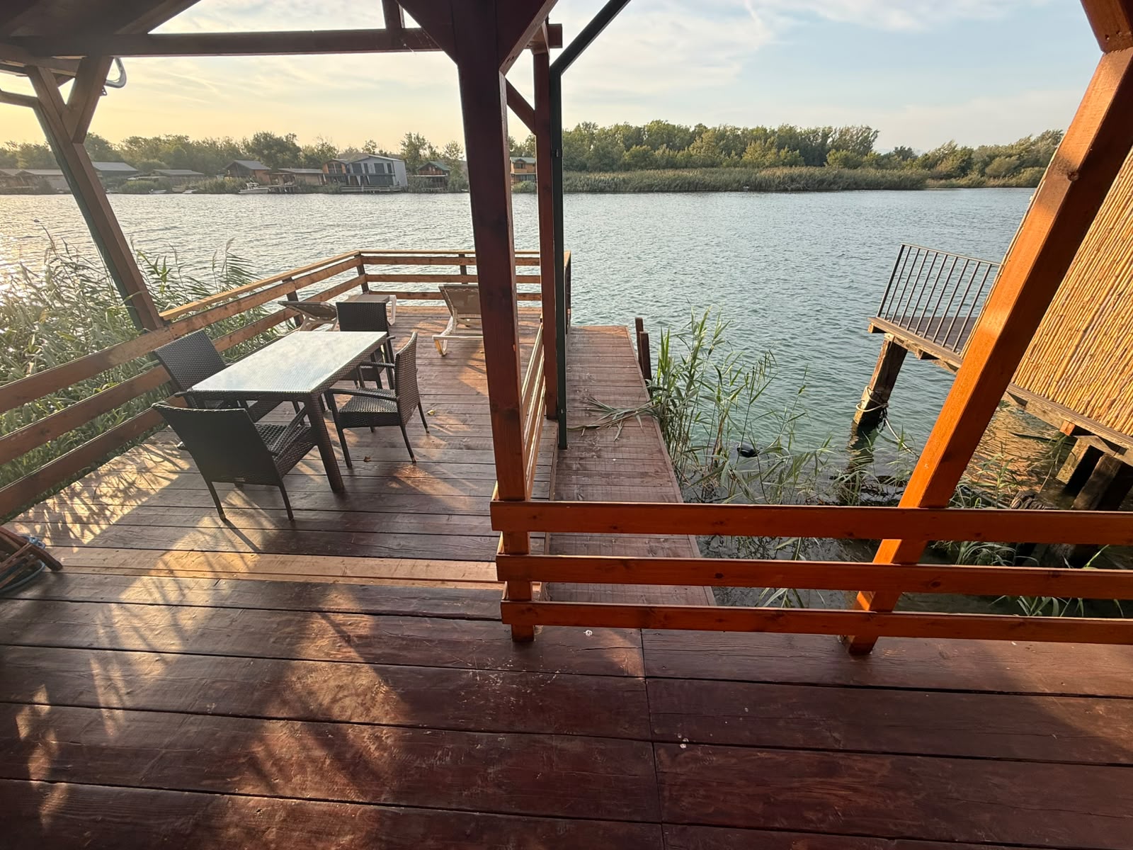 Experience serene riverside living in Ulcinj, Montenegro with this exceptional property featuring a spacious wooden deck complete with elegant table, comfortable chairs, and secure railings for relaxing or entertaining guests. Overlooking a tranquil river bordered by lush reeds, the private dock on the right offers easy water access—perfect for boating enthusiasts or nature lovers. The breathtaking views include mature trees and charming homes set against a clear sky, creating an inviting atmosphere ideal for investment or your dream home. Discover prime real estate opportunities on montenegro-listings.me, a trusted project of ekosphere doo in Ulcinj—a gateway to Montenegro’s vibrant property market.