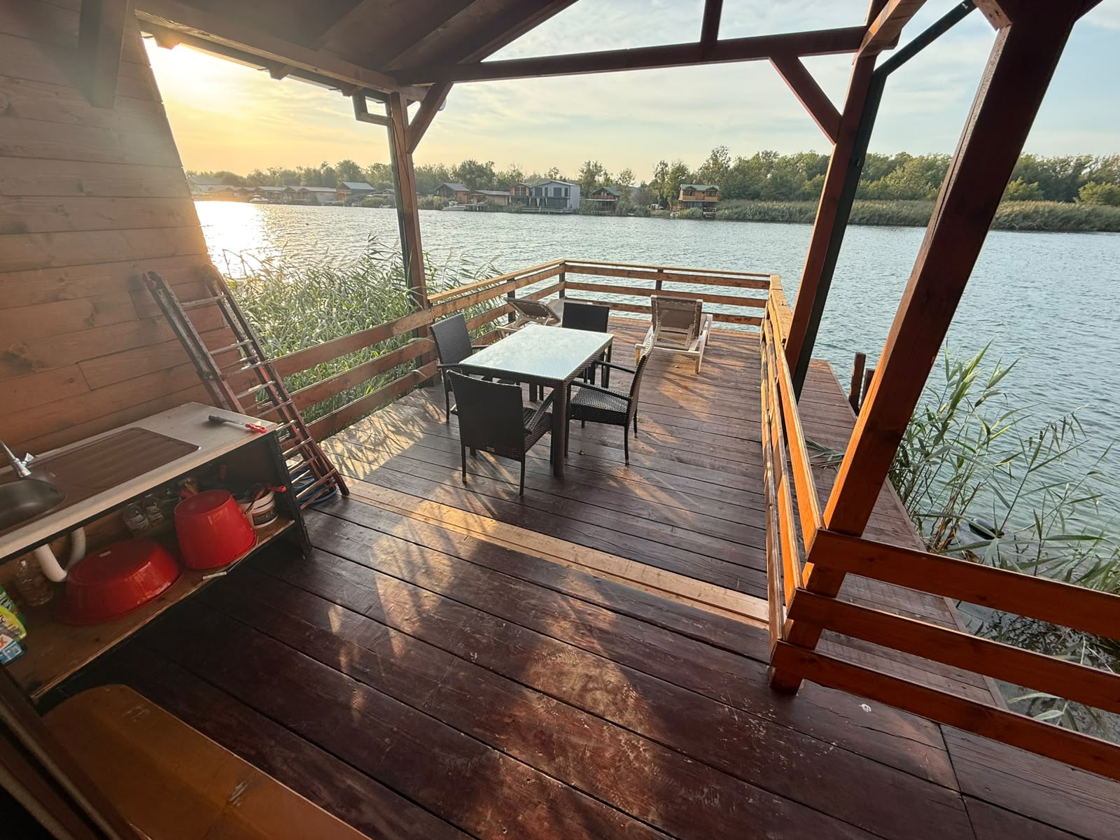Experience the ultimate in riverside living with this exclusive property for sale in Ulcinj, Montenegro, featured on montenegro-listings.me—a project of ekosphere doo. This spacious wooden deck overlooks a serene river and boasts breathtaking panoramic views of the water and charming distant houses under a clear open sky. Perfect for entertaining or relaxing, the deck is thoughtfully designed with a modern outdoor kitchen equipped with sink and gas tanks, a stylish dining table for four, and two luxurious reclining loungers. Ideal for investors or homebuyers seeking prime waterfront real estate in Montenegro’s thriving property market, this unique opportunity combines comfort, luxury amenities, and an unbeatable location for tranquil living or lucrative rental returns.