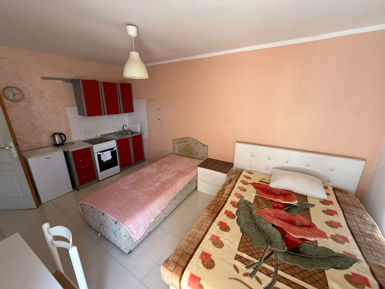 Charming Ulcinj studio apartment for sale, featuring light pink walls, a cozy single bed and twin bed setup, elegant white nightstands, and a modern compact kitchen with striking red cabinets, stove, sink, and kettle. This well-designed space also includes a wall clock positioned by a pristine white door—perfect for property investors seeking efficient layouts in the Montenegro real estate market. Discover exceptional value with this move-in-ready apartment exclusively on montenegro-listings.me—a trusted project of ekosphere doo in Ulcinj.