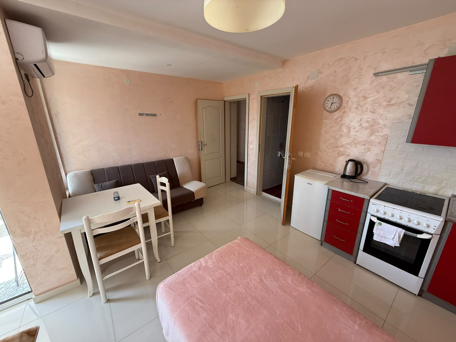 Discover an excellent investment opportunity with this charming, fully furnished apartment in Ulcinj, Montenegro, available exclusively through montenegro-listings.me by ekosphere doo. The thoughtfully designed space features a comfortable bed, modern dining table with chairs, cozy sofa, and a practical kitchenette equipped with a stove, fridge, and kettle—perfect for daily living or lucrative rental income. Stylish light pink walls and bedding create an inviting atmosphere, complemented by durable tiled flooring throughout. The layout includes two access doors leading to additional rooms for versatile use of space. Ideal for investors seeking property in the thriving Ulcinj real estate market or buyers looking to secure a tranquil Montenegrin retreat close to Adriatic beaches.