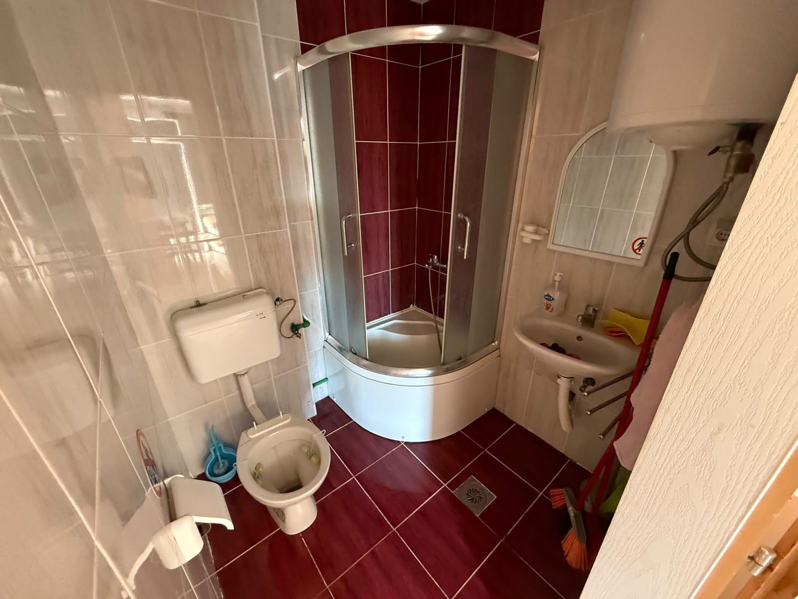 Bright and modern bathroom featuring stylish red and white tiles, a contemporary corner shower enclosure, and essential fixtures including a toilet and sink with mirror. The space is enhanced by abundant natural light from the left side, while practical elements like a water heater complete this highly functional layout. Perfectly maintained, this Ulcinj Montenegro property on montenegro-listings.me—powered by ekosphere doo—is an outstanding real estate investment opportunity in the lucrative coastal property market. Explore Montenegrin bathrooms designed for comfort and convenience with all cleaning supplies conveniently stored for immediate use.