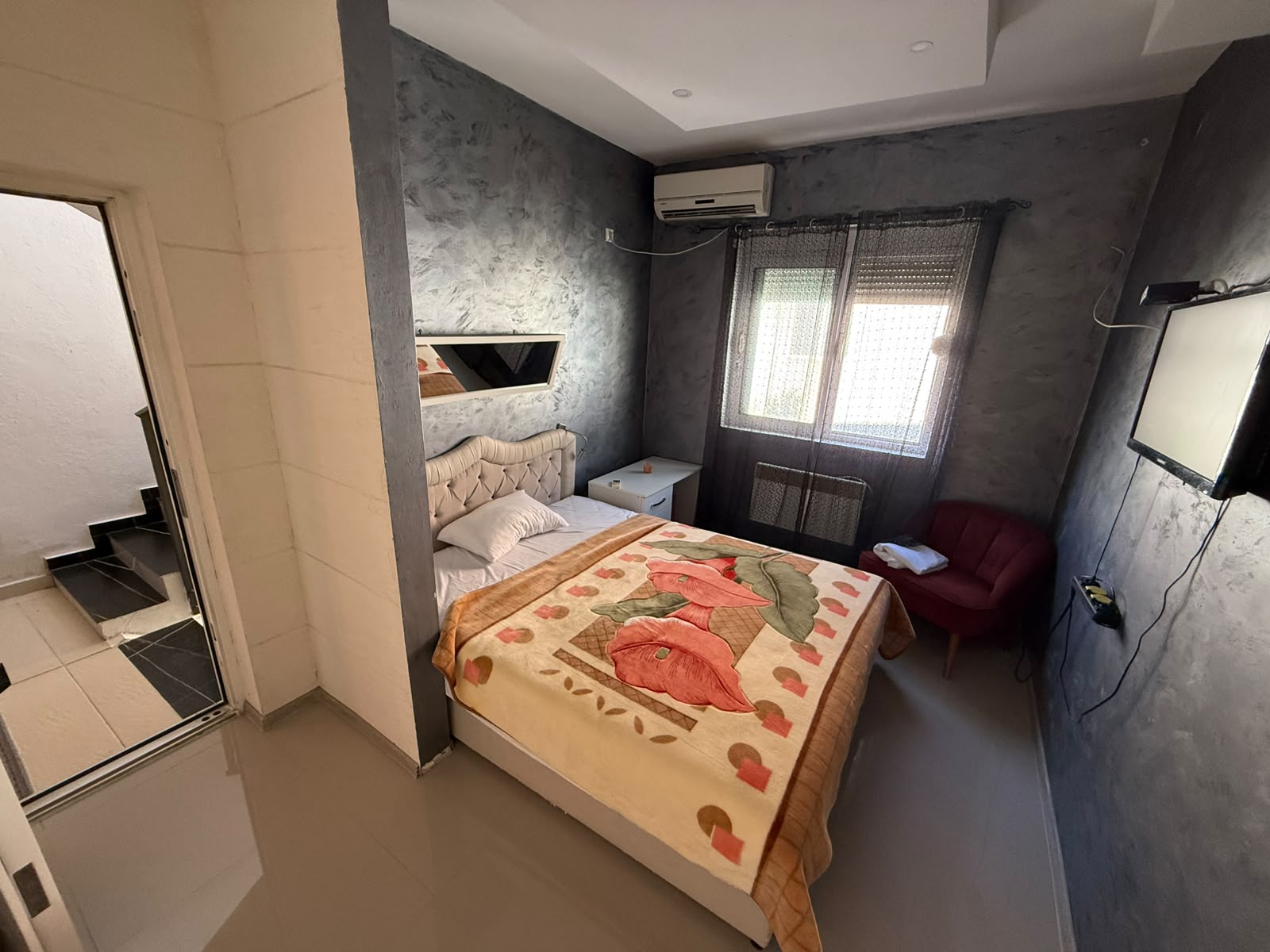 Charming small bedroom for sale in Ulcinj, Montenegro—ideal for investors or as a cozy holiday retreat. This inviting space features a comfortable bed adorned with a stylish floral blanket, enhancing both comfort and aesthetic appeal. Amenities include a modern wall-mounted TV, efficient air conditioning above the window for year-round climate control, and thoughtful furnishings such as a red accent chair and nightstand. The convenient doorway leads directly to the hallway and stairs, offering easy access throughout the property. Explore prime Ulcinj real estate listings on montenegro-listings.me, presented by ekosphere doo—your trusted partner in property investments in Montenegro’s thriving market.