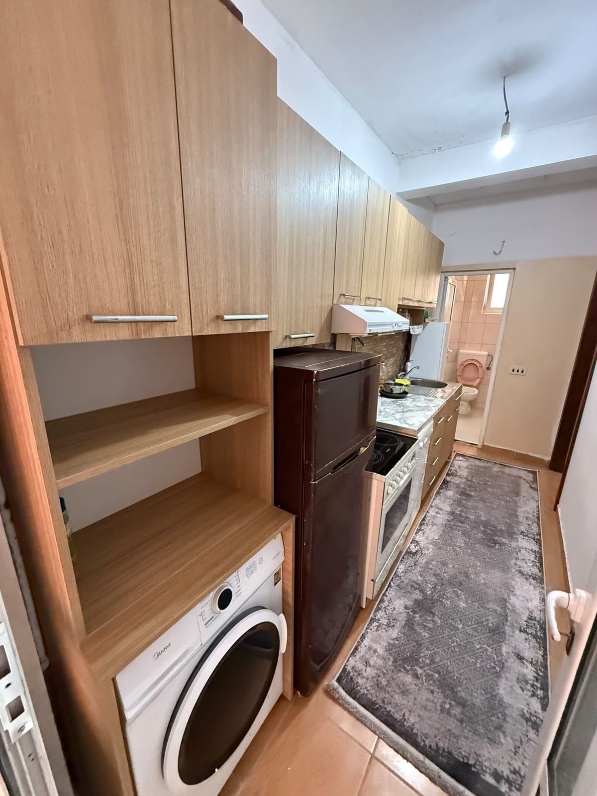 Discover a functional, well-equipped kitchen in Ulcinj, Montenegro, ideal for residential living or investment. Featuring quality wooden cabinetry, modern appliances—including a washing machine, refrigerator, stove, and oven—all conveniently arranged along one wall for optimal space efficiency. A sleek gray rug enhances the inviting atmosphere, while a door at the end of the kitchen opens directly to a contemporary bathroom with visible sanitary fittings. This property is showcased on montenegro-listings.me—a premier real estate platform from ekosphere doo in Ulcinj—making it an excellent opportunity for buyers seeking stylish homes or lucrative rental assets in the thriving Montenegro property market.
