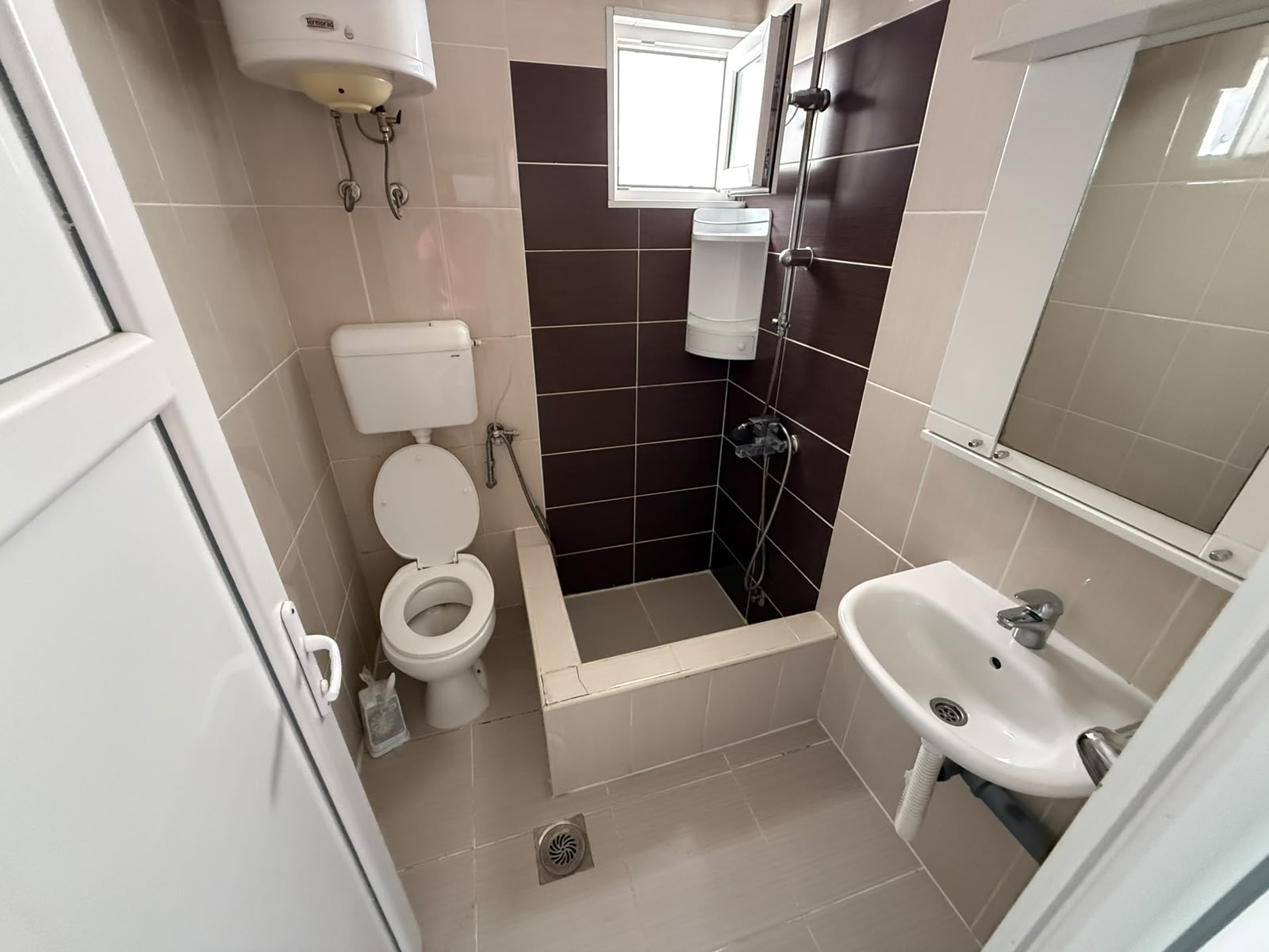 Modern, fully-tiled bathroom in Ulcinj, Montenegro, ideal for property investors and home buyers seeking quality real estate. This contemporary space features a toilet, wall-mounted sink, spacious open shower area with central floor drain (no curtain for a sleek look), and two efficient water heaters—perfect for year-round comfort. The elegant beige and brown tiled walls add warmth and sophistication, while natural light pours in through the window above the mirror. Discover high-potential investment opportunities like this on montenegro-listings.me—a project by ekosphere doo Ulcinj—your trusted partner in Montenegro’s dynamic property market.