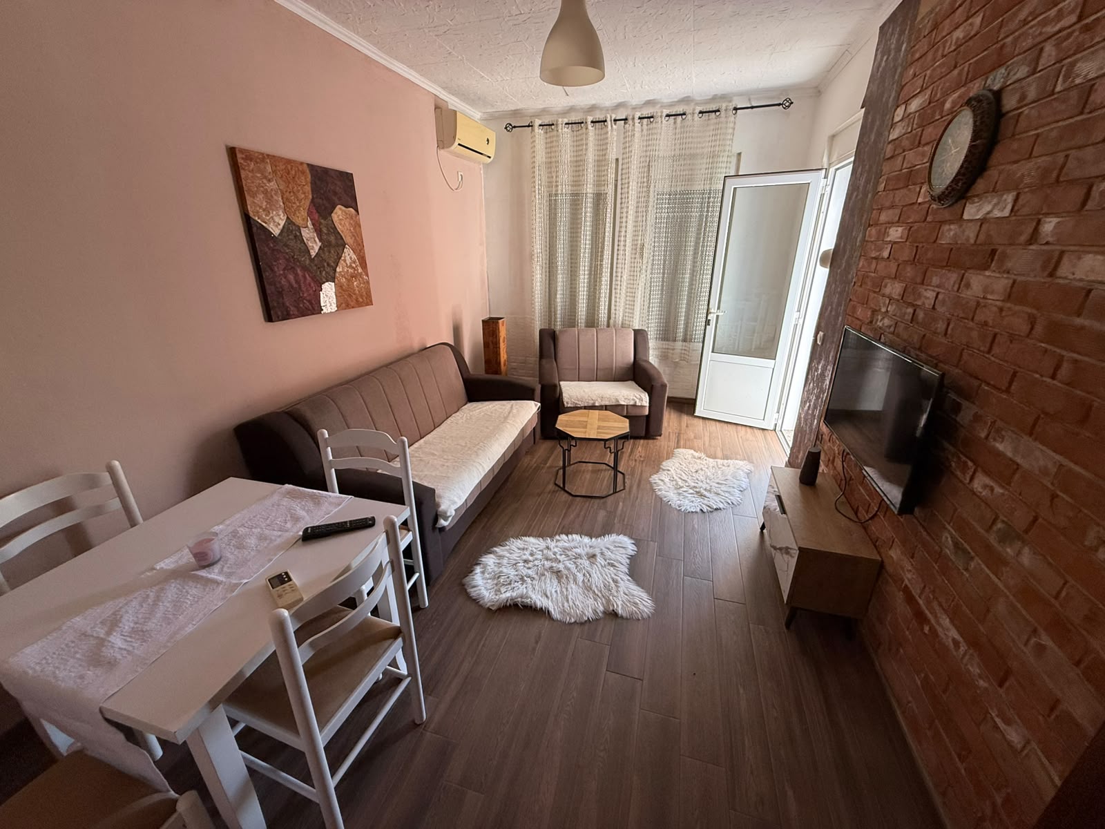 Bright and inviting living room in Ulcinj, Montenegro, featuring a stylish brown sofa and armchair, modern coffee table, flat-screen TV set against an elegant brick accent wall, and a spacious dining area with a contemporary table and four chairs. Sunlight streams through sheer curtains on the patio door, illuminating two plush white rugs for added comfort. This turnkey property is the perfect investment opportunity in Ulcinj’s thriving real estate market. Find your ideal home or profitable rental at montenegro-listings.me—an exclusive project by ekosphere doo in Ulcinj.