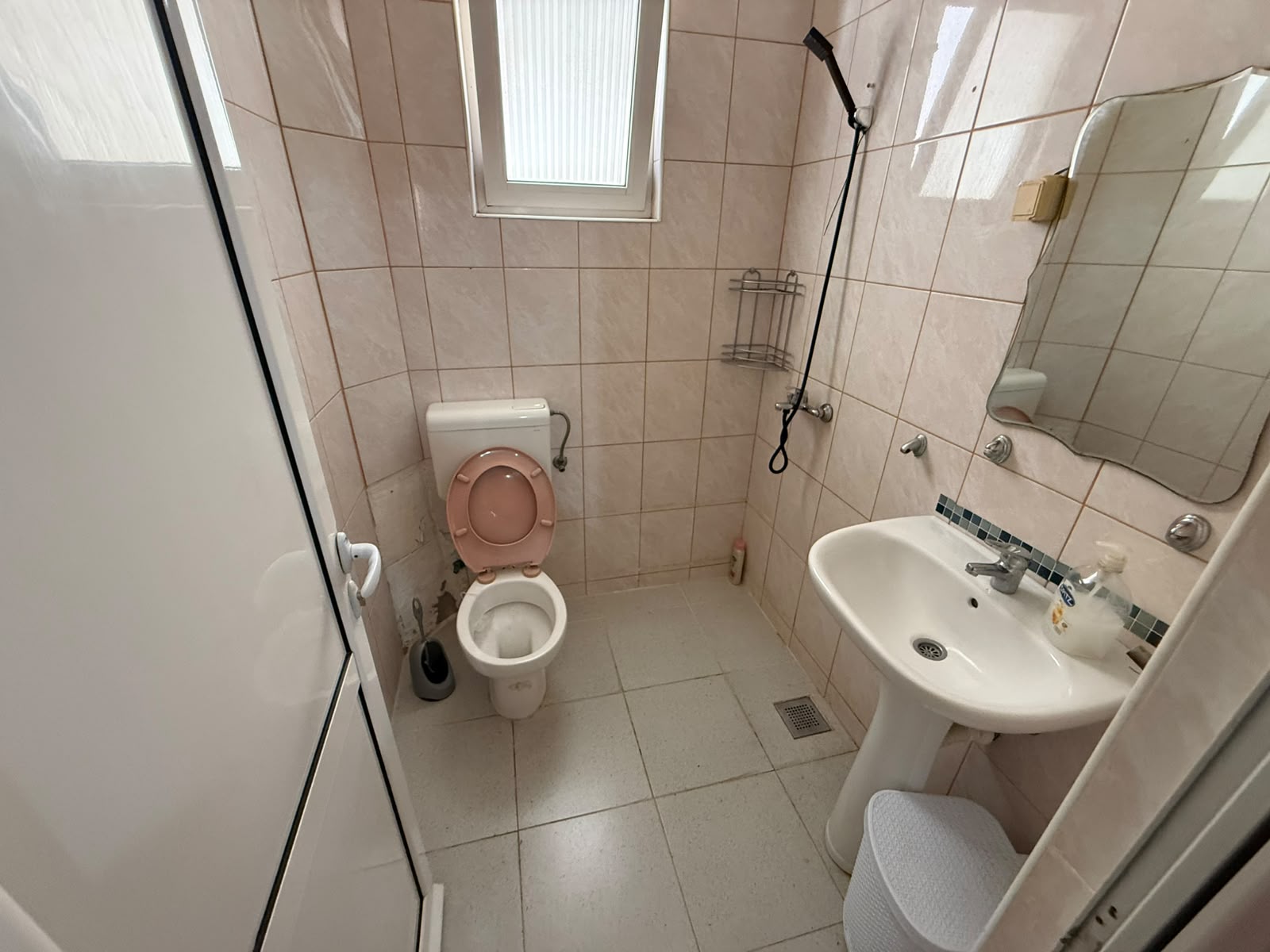 Discover this charming, efficiently designed bathroom in Ulcinj, Montenegro – an ideal investment opportunity presented by montenegro-listings.me, a project of ekosphere doo. The space features modern light-colored tile flooring, a well-maintained toilet with an attractive pink seat, stylish sink and mirror setup, practical shelving for storage, and natural light from the window. Additional amenities include a quality showerhead, convenient soap dispenser, toilet brush set, and discrete trash can. Perfect for buyers seeking functional living or rental potential in the vibrant Ulcinj property market.