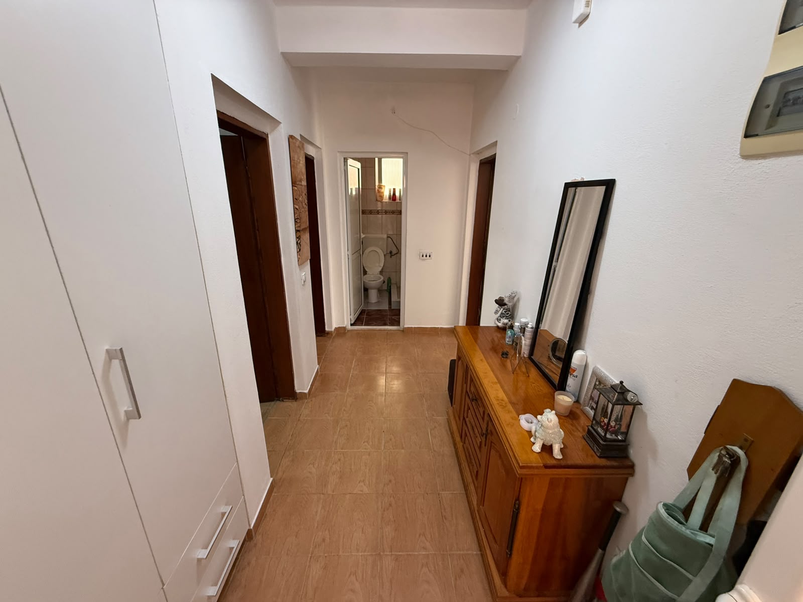 Spacious and inviting hallway in an Ulcinj property, featuring stylish light brown tile flooring, crisp white walls, a classic wooden dresser with an elegant mirror on the right, and functional cabinetry on the left—enhancing storage options. This well-lit corridor offers access to three separate rooms and leads to a modern bathroom with visible toilet facilities. Ideal for investors seeking prime real estate in Montenegro, this home exemplifies quality construction and practical design. Discover standout properties like this through montenegro-listings.me—your trusted partner for Ulcinj real estate opportunities and a dedicated project of ekosphere doo.