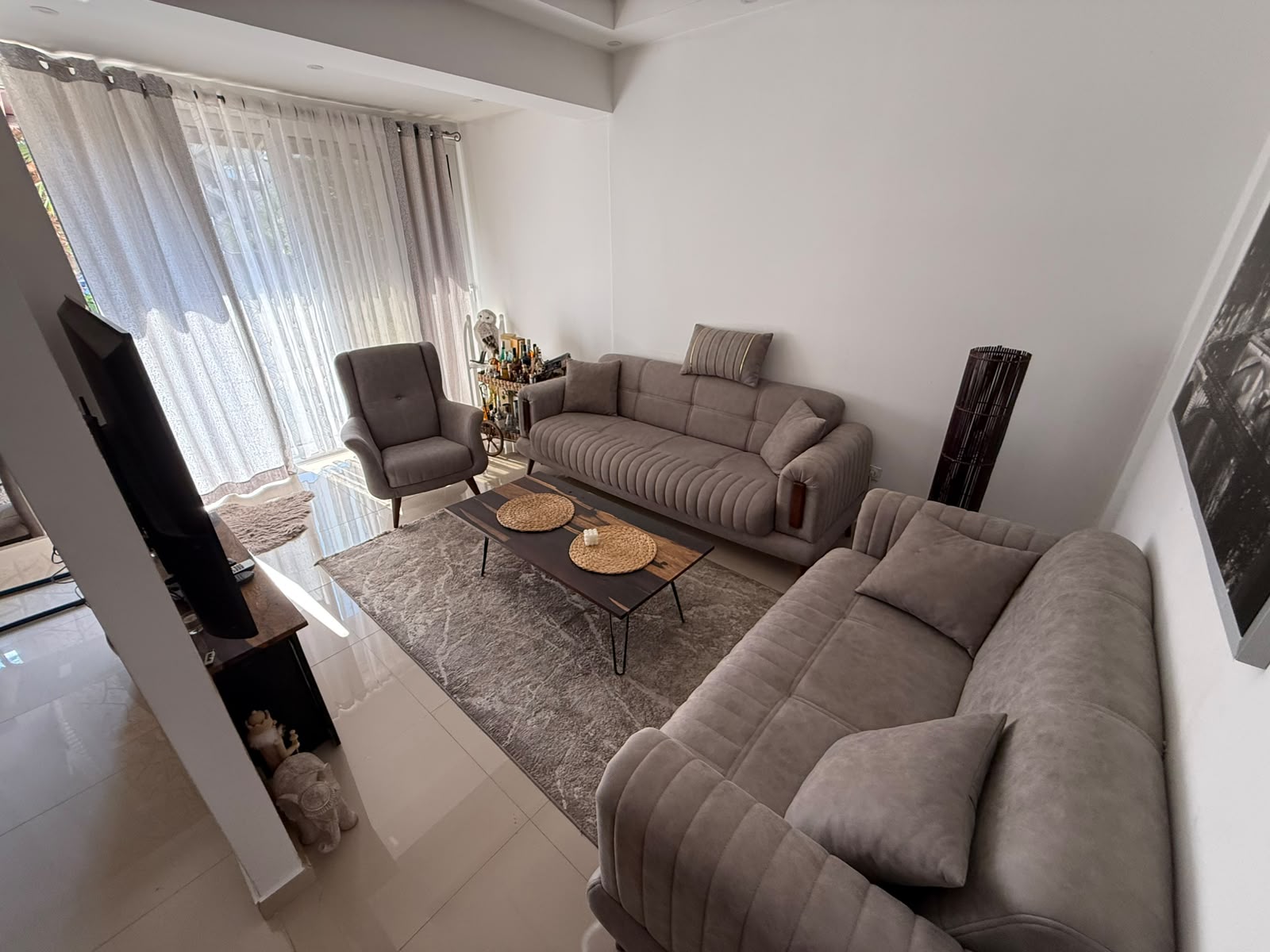 Elegant and bright living room in Ulcinj, Montenegro, featuring two contemporary gray sofas, a stylish armchair, a premium wooden coffee table with woven placemats, modern area rug, flat-screen TV, chic bar cart, and floor lamp. Expansive sheer white curtains allow abundant natural sunlight to fill the space against light-colored walls and flooring—creating an inviting ambiance ideal for entertaining or relaxing. This turnkey property is an attractive investment opportunity in the Montenegro real estate market. Discover more exclusive homes through montenegro-listings.me—a project of ekosphere doo in Ulcinj.