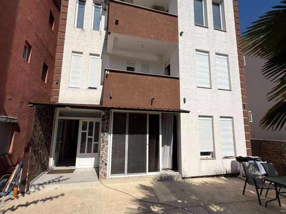 Modern three-story residential building for sale in Ulcinj, Montenegro, featuring elegant white and brown exterior finishes. The property offers spacious balconies on the second and third floors, expansive windows for abundant natural light, and a stylish glass patio door connecting to a charming outdoor seating area on a paved terrace. Located under clear blue skies in a desirable Montenegrin neighborhood, this listing is an exceptional investment opportunity in the fast-growing Ulcinj real estate market. Discover more exclusive properties at montenegro-listings.me—a project by ekosphere doo in Ulcinj—connecting you to premium Montenegro investments.
